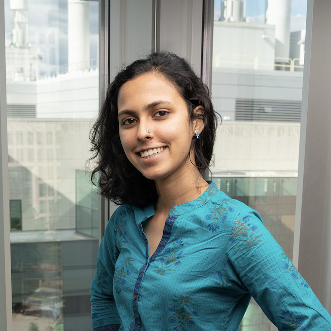 Image contains one (1) Rujul smiling and a fraction of the Stata center (MIT Bldg 32)